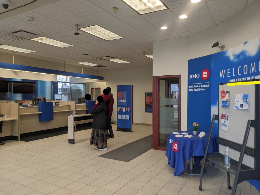 BMO Bank branch with a lien of people, one teller open only, and a sign that says "How can we help you?"